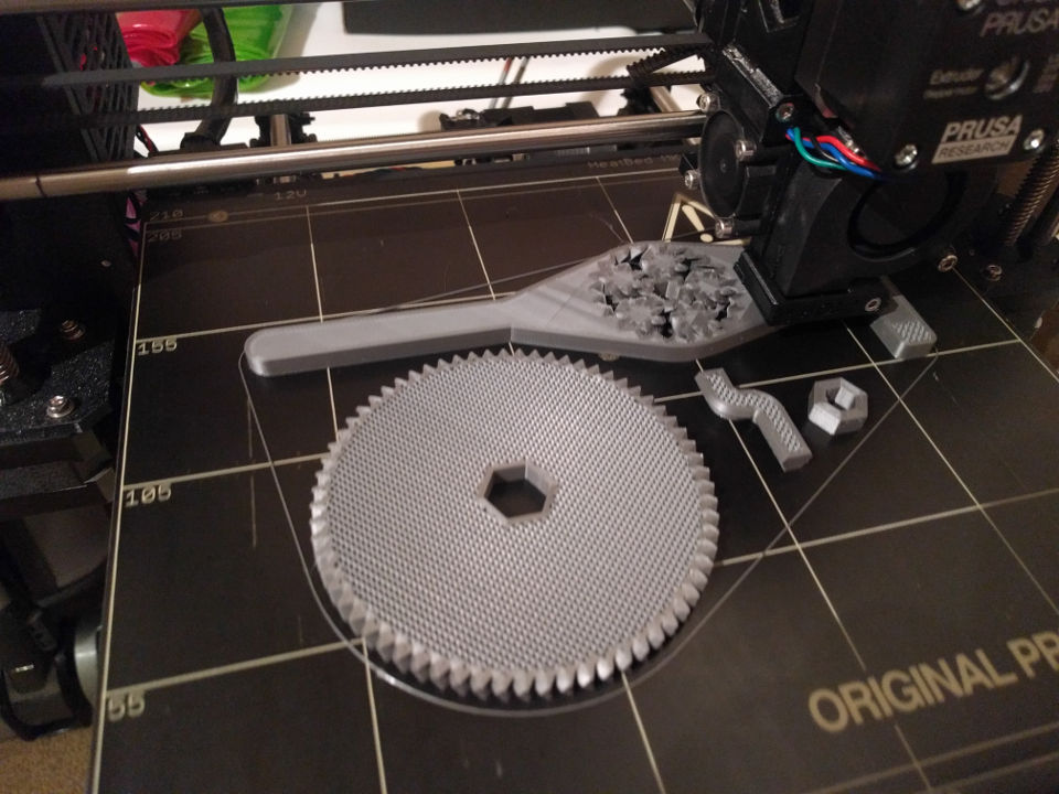 A large gear, a leg, a nut, and a bent shaft on the bed of a 3d-printer. The print is in progress; the gear and shaft's dense inner infill has not yet been covered.